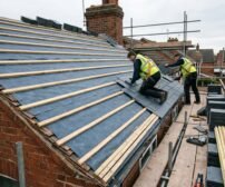Roofers on scaffolding during a roof replacement, showing the stages of laying underlay, timber battens, and new slate tiles.
